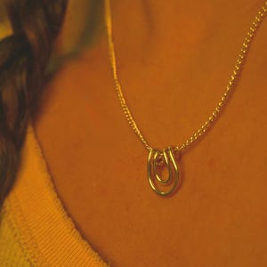 May include: A gold-colored necklace with a double-loop pendant. The chain is delicate, and the pendant is a simple, elegant design. The necklace is worn against a yellow shirt.