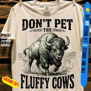 May include: A cream-colored t-shirt featuring a black graphic illustration of a bison in a dusty landscape. The text "DON'T PET THE FLUFFY COWS" is printed in a bold, dark font above and below the central animal image on the front of the shirt.