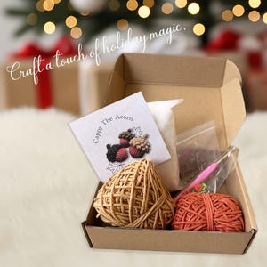 May include: Open cardboard box containing craft supplies. Includes two balls of yarn, one tan and one orange, a crochet hook, and a small booklet with the text "Cappy The Acorn". The text "Craft a touch of holiday magic" is written above the box.