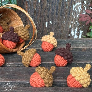 May include: Crocheted acorns in various shades of brown and orange are scattered on a wooden surface, with some spilling from a woven basket. The acorns have textured caps and rounded bodies, evoking a rustic, autumnal theme.