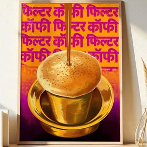 May include: A framed art print featuring a golden coffee filter and cup, with coffee being poured into the cup. The background is orange with repeating pink text. The print is displayed on a wooden surface.