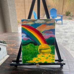 May include: A colorful painting of a rainbow arching over a green field with a pot of gold. The artwork is displayed on a black easel, with a background of a patio and blue chairs.