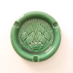 May include: Green ceramic ashtray with a raised design of crossed flags and the text "Allied Air Forces Central Europe".