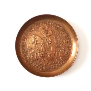 May include: A round copper tray with an embossed design of a person riding a horse. The design is surrounded by a decorative border.