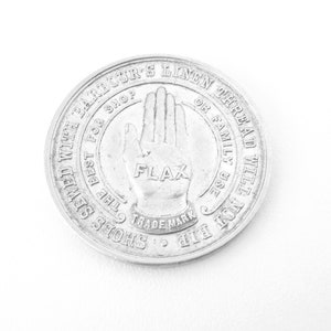 May include: A silver metal token with the text "Barbour's Linen Thread, The Best For Shop or Family Use, Rip Shoes Sewed With Flax, Trade Mark" around the edge. A hand with the word "Flax" on the palm is in the center.