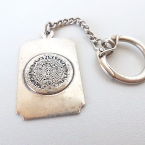 May include: Silver keychain featuring a circular design with intricate details, resembling an ancient calendar. The keychain has a silver ring for attaching keys.