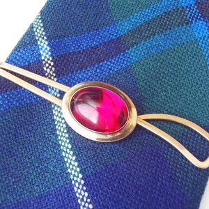 May include: A gold tie clip with a red oval-shaped gemstone. The clip is on a plaid fabric background.
