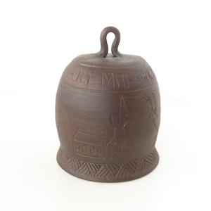 May include: A brown ceramic bell with a loop handle. The bell is decorated with carved designs, including a church, houses, and trees. The text "ERRY MOHAWK" is carved around the top of the bell.
