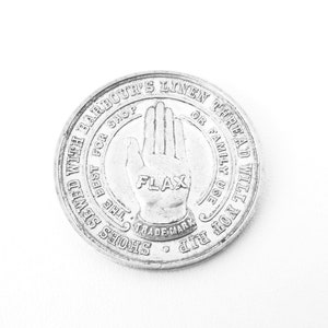 May include: A silver token with the text "SHOES SEWED WITH BARBOUR'S LINEN THREAD WILL NOT RIP FOR FAMILY USE OR SHOP THE BEST FOR FLAX TRADE MARK" around the edge. A hand with the word "FLAX" on the palm is in the center.