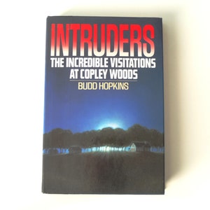 May include: A book cover with a dark blue background and a white house in the distance. The title "Intruders" is in red and white letters. The subtitle is "The Incredible Visitations at Copley Woods" and the author is "Budd Hopkins".