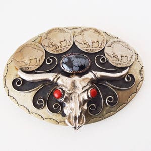 May include: A silver belt buckle with a black and silver bull skull design. The skull has red stones for eyes and a black stone in the center of the forehead. The buckle is decorated with five silver buffalo nickel coins.