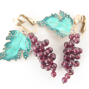 May include: A pair of clip-on earrings featuring a green enamel grape leaf and a cluster of dark red glass beads. The earrings are made of gold-tone metal.