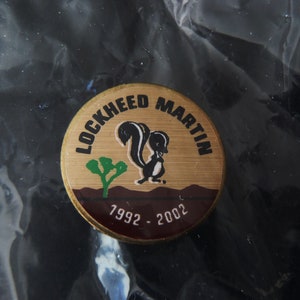 May include: A gold-colored round pin with the text "Lockheed Martin" and a black and white skunk illustration. The pin also has the text "1992 - 2002" on it.