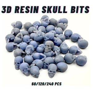 Op de afbeelding: Een verzameling kleine, blauwe hars schedel bits. De schedels zijn in verschillende houdingen en maten, met zichtbare details. Tekst bovenaan luidt "3D RESIN SKULL BITS" en onderaan "60/120/240 PCS".