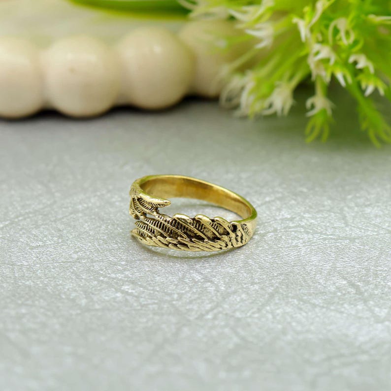 May include: A gold ring with a detailed feather design. The ring is made of a shiny, metallic material and features intricate patterns that resemble feathers. The ring is a jewelry item, suitable for everyday wear or special occasions.