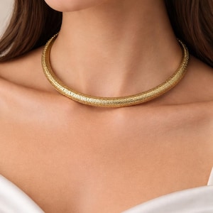 Puede incluir: Un collar dorado con superficie texturizada. El collar es una banda gruesa y redondeada que se ajusta al cuello. La joya la lleva una persona con pelo largo castaño y una blusa blanca.