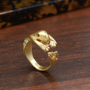 Puede incluir: Un anillo de rana de color dorado. El anillo presenta un diseño detallado de rana con brazos y piernas extendidos. El anillo está abierto en la parte inferior, lo que permite ajustar la talla. Los ojos y la textura de la piel de la rana son visibles.