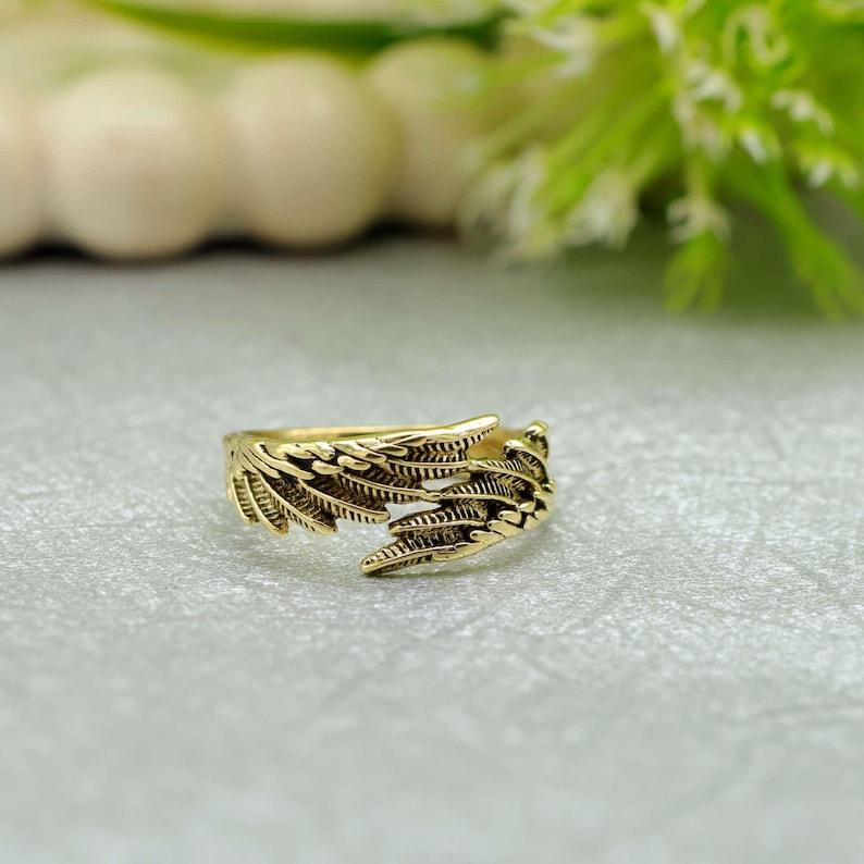 May include: A gold-coloured ring with a detailed feather design. The ring features two stylised feather wings that wrap around the finger. The ring is made of a shiny, metallic material and is set against a soft, neutral background.