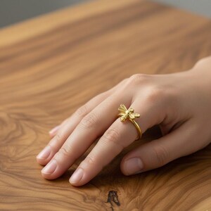 Pode incluir: Anel dourado com um design de libélula. O anel é usado em um dedo. A libélula tem asas e corpo detalhados. O fundo é de madeira.