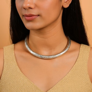 Puede incluir: Un collar plateado grabado. El collar es un tubo grueso y redondeado con un patrón floral detallado. El collar se usa alrededor del cuello, descansando sobre la clavícula. El fondo es de color naranja claro sólido.
