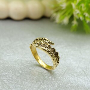 May include: A gold-coloured ring with a feather design. The ring features detailed feather patterns that wrap around the band. The ring is set against a soft, neutral background, highlighting its intricate design.