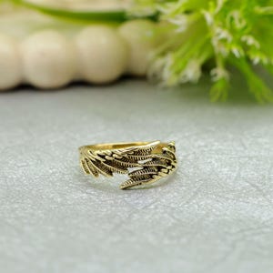 May include: A gold ring with a detailed feather design. The ring has intricate detailing that mimics the texture of feathers. The ring is set against a soft, neutral background, highlighting its metallic sheen and craftsmanship.