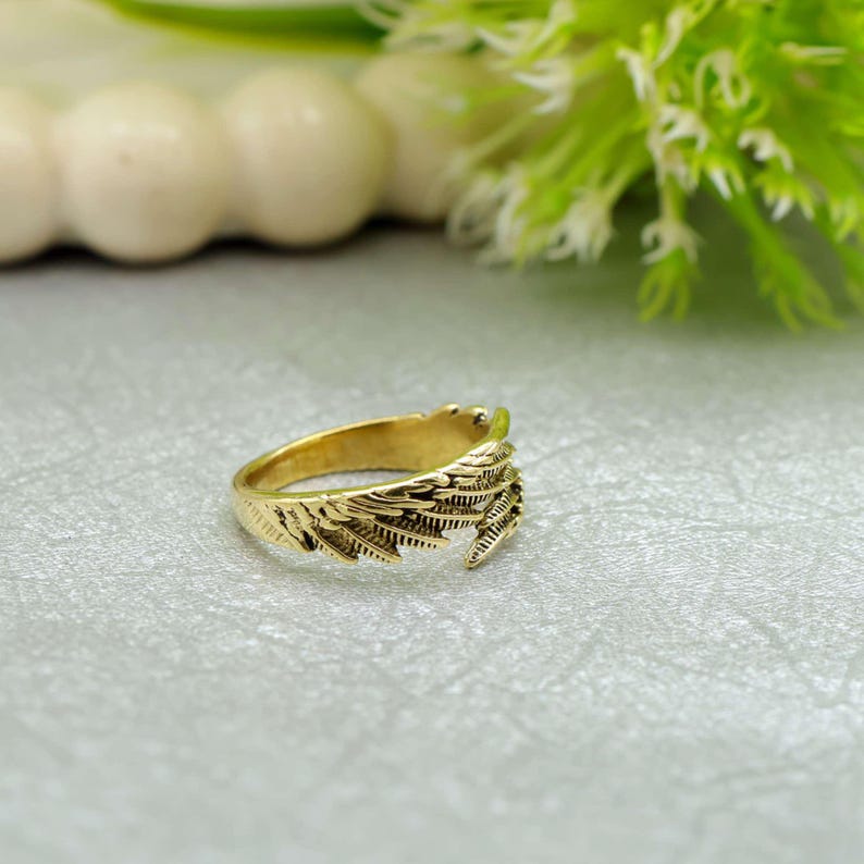May include: A gold-coloured ring featuring a detailed feather design. The ring is made of a shiny, metallic material and is set against a soft, neutral background. The feather design wraps around the ring, creating a unique and elegant look.
