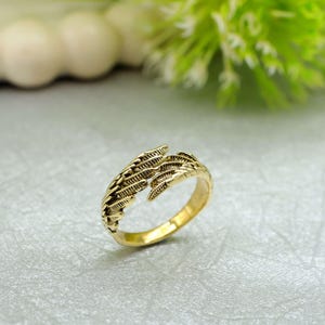 May include: A gold-coloured ring with a detailed feather design. The ring has an open design with two feather-like elements that wrap around the finger. The ring is set against a light grey surface with green foliage in the background.