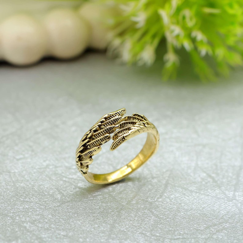 May include: A gold-coloured ring with a detailed feather design. The ring has an open design with two feather-like elements that wrap around the finger. The ring is set against a light grey surface with green foliage in the background.