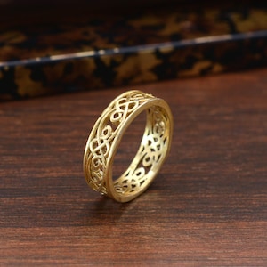 Può includere: Un anello color oro con un intricato design a nodo celtico. L'anello presenta un motivo traforato, con il design ripetuto attorno alla fascia. L'anello è probabilmente in metallo e viene esposto su una superficie di legno.