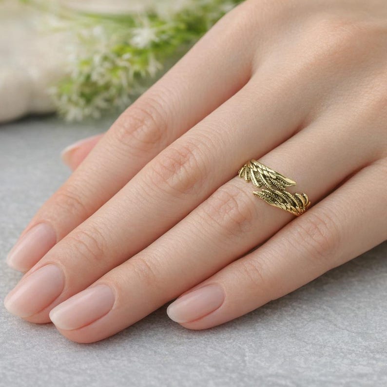 May include: A gold-coloured ring with a leaf design is worn on a finger. The ring wraps around the finger with two detailed leaf-like structures. The ring is made of a shiny, metallic material.