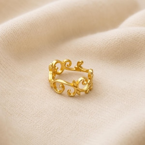 May include: A gold-toned ring featuring an openwork design composed of repeating swirling scroll and leaf-like motifs. The band has a polished metallic finish and a continuous pattern that wraps around the entire circumference of the circular ring.