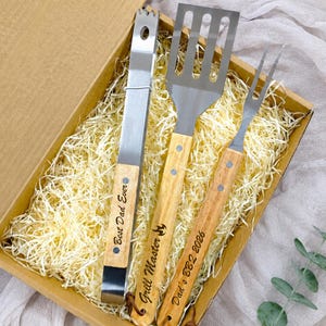 Peut inclure: Un ensemble de trois ustensiles de barbecue en acier inoxydable avec des manches en bois clair, comprenant une pince, une spatule fendue et une fourchette à deux dents, présentés dans une boîte cadeau en carton remplie de papier déchiqueté.