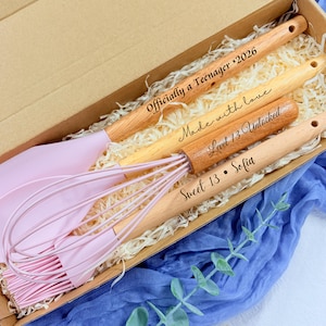 Könnte beinhalten: Geschenkbox mit rosa Silikon- und Holz-Küchenutensilien. Die Holzgriffe sind mit Sätzen wie "Officially a Teenager 2026" und "Sweet 13 • Sofia" graviert. Die Box ist mit Papier gefüllt.