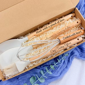 Può includere: Un set di utensili da cucina in scatola, con una spatola, una frusta, un pennello e cucchiai di legno. I manici in legno sono incisi con frasi come "Made with a pinch of love" e "Cooked with heart". Il set è presentato in una scatola di cartone.