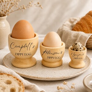 May include: Three personalised wooden egg cups, each holding a different egg: a brown chicken egg, a pale chicken egg, and a speckled quail egg. The cups are engraved with names and the words "Dippy Egg".