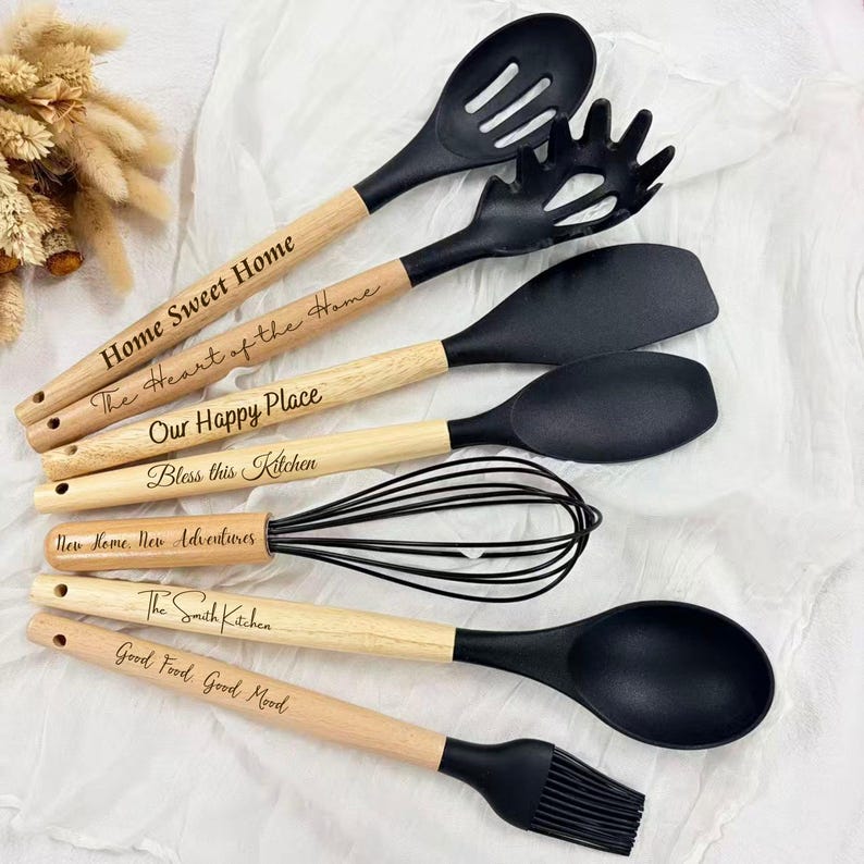 May include: A collection of black kitchen utensils with wooden handles. The utensils include a slotted spoon, spaghetti fork, spatula, solid spoon, whisk, and basting brush. Text is engraved on the handles, including phrases like "Home Sweet Home" and "Bless this Kitchen."