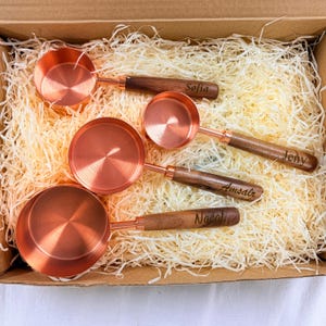 May include: Four copper measuring cups with wooden handles, each engraved with a name: Sofia, Jeny, Amsalr, and Nocol. The cups are nested in a cardboard box filled with straw-like packing material. The copper has a shiny, reflective surface.