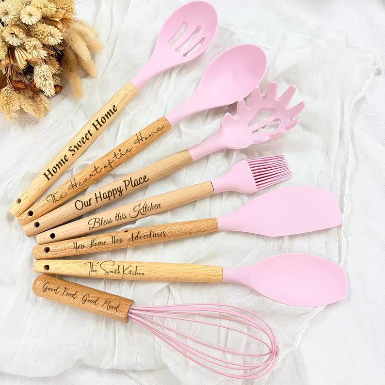 May include: A set of pink silicone kitchen utensils with wooden handles. The handles are engraved with phrases like "Home Sweet Home" and "Good Food, Good Mood." Includes a slotted spoon, spatula, whisk, and more. Ideal for cooking and baking.