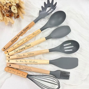 May include: A collection of gray and wood-handled kitchen utensils, including a whisk, spatula, and slotted spoon. Each utensil has a wooden handle with engraved text such as "Home Sweet Home" and "Bless this Kitchen". The utensils are arranged on a white surface.