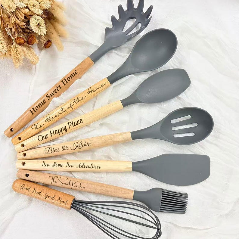 May include: A collection of gray and wood-handled kitchen utensils, including a whisk, spatula, and slotted spoon. Each utensil has a wooden handle with engraved text such as "Home Sweet Home" and "Bless this Kitchen". The utensils are arranged on a white surface.