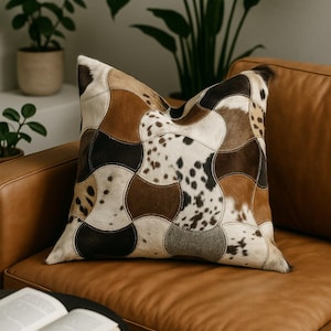Op de afbeelding: Een decoratief vierkant kussen met een patchwork ontwerp. Het heeft verschillende tinten bruin, wit en zwart koeienleer stukken aan elkaar gestikt. Het kussen ligt op een bruine leren bank.