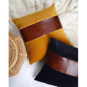 Puede incluir: Dos cojines cuadrados con una banda horizontal central de cuero marrón. Un cojín es de terciopelo amarillo mostaza y el otro es de terciopelo negro. Ambos cojines tienen una textura lisa en el cuero y un acabado suave en la tela.
