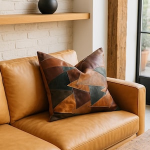 May include: A patchwork cushion in various shades of brown and green sits on a tan leather sofa. The cushion's geometric design complements the sofa's warm tones. The sofa is next to a wooden table, creating a cosy living space.