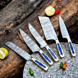 Pode incluir: Um conjunto de cinco facas de aço Damasco com lâminas estampadas e cabos azuis e dourados. As facas estão dispostas sobre uma superfície de madeira com metade de limão e pequenos pimentões.