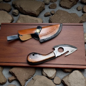 May include: Two knives on a wooden surface. One is a hunting knife with a brown leather sheath. The other is a small, curved knife with a wooden handle and a hole near the blade. The knives are displayed on a brown wooden tray.
