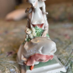 May include: A porcelain figurine depicting a woman carrying a child on her back. The woman wears a white dress with a red sash and holds flowers. The figurine is on a rectangular base and has a delicate, hand-painted appearance.