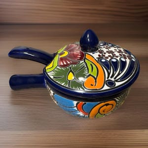 May include: A hand-painted ceramic casserole dish with a lid. The dish features a vibrant floral design in shades of blue, green, orange, and red. It has two blue handles and a blue knob on the lid. The dish is likely used for serving food.