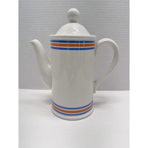 May include: A white ceramic coffee pot featuring a tall cylindrical body, a curved handle, and a spout. The pot includes a removable lid with a spherical knob. Horizontal orange and blue stripes decorate the upper and lower sections of the pot.
