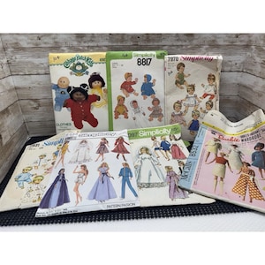 May include: A collection of seven vintage sewing patterns for dolls, including Cabbage Patch Kids, Simplicity, and McCall's brands. The paper envelopes feature illustrations of various doll outfits, including dresses, jumpsuits, and pajamas, with printed text identifying pattern numbers and sizes.